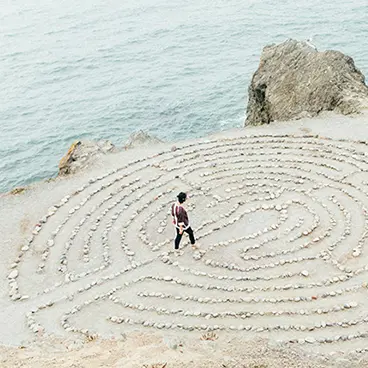 aerial view of a woman walking through a spiraled labyrinth made out of stones on a beach overlooking the ocean