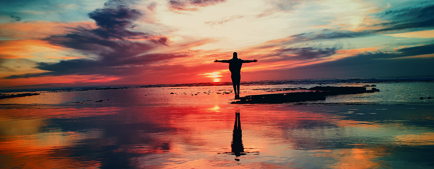 multicolored sunset by the ocean with a person standing with their arms outstretched