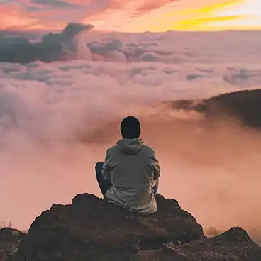 man sitting on a mountain looking out at clouds during sunset